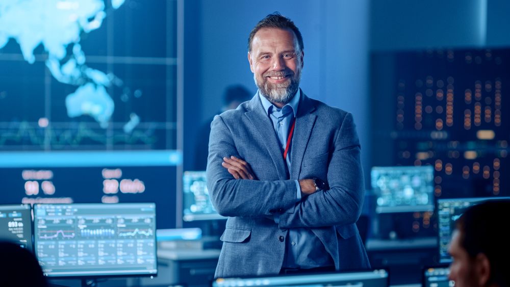 Confident Executive Stands in High-tech Control Room. Team Monitors Global Data on Screens. Blue-lit Environment With World Map Displays. Advanced Technology for Real-time Analysis.