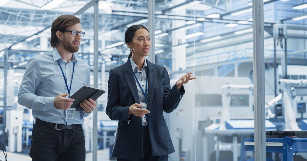 Two Diverse Engineers Walking in Office, Using Tablet Computer at a Modern Electronics Factory. Female and Male Specialists Chatting, Monitoring Robots on Modern Assembly Line.