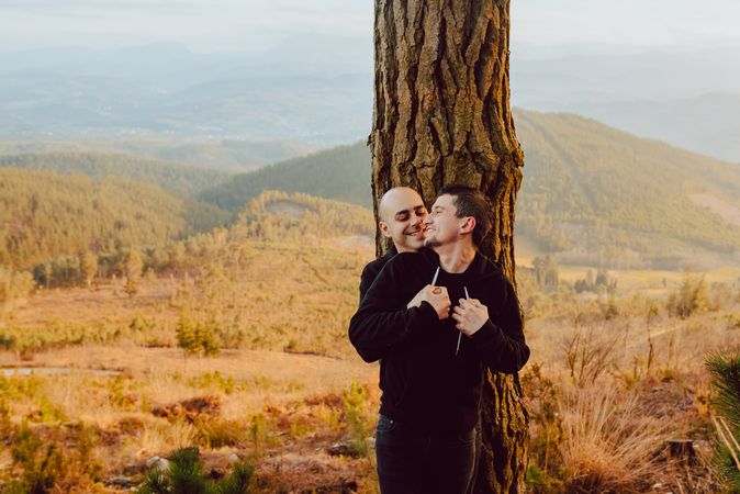 Smiling men hugging near tree in forest on hill