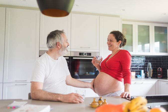 Pregnant woman with glass of water as her husband touches her stomach in kitchen