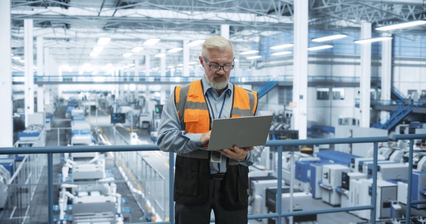 Engineer Standing on a Platform, Using Laptop Computer at an Electronics Factory. Machines are Undergoing Maintenance, Specialist Monitoring the Updates Through Online Software