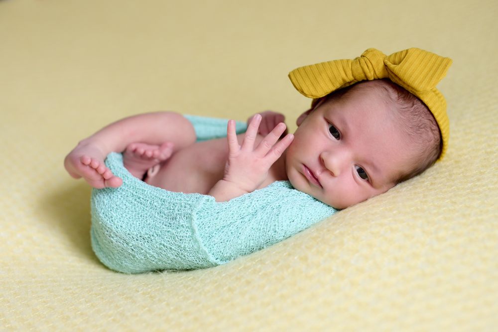 Close-up of a newborn baby girl, inside a ball of blue wool on a yellow blanket with a yellow bow on her head