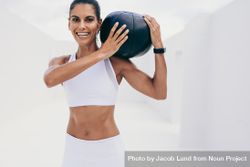 Smiling woman in fitness wear doing workout with a medicine ball on her shoulder 48ajjb