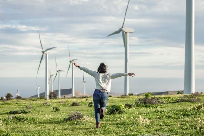 Back view of excited woman in light sweater and denim pants standing beside wind turbines