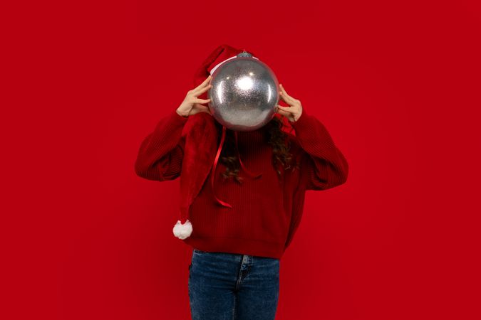 Girl in Santa hat playfully covers face with Christmas ornament.