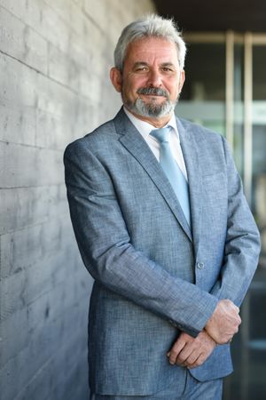 Grey haired man in formal suit smiling next to grey brick wall