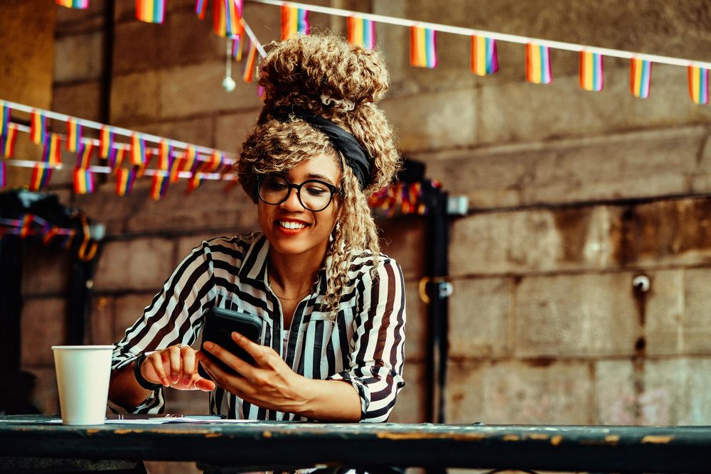 Happy Black woman in stripe shirt texting on phone in cafe