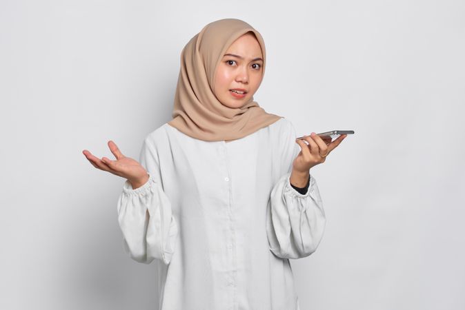 Asian Muslim woman smiling while with arms up in confusion using phone speaker phone
