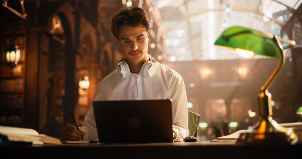 Concentrated Young Man Studying in a Classical Library, Using Laptop and Headphones for E-Learning.