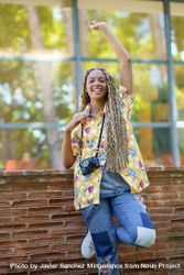 Female in bold patterned shirt leaning on brick wall with camera and arm above head 478kgb
