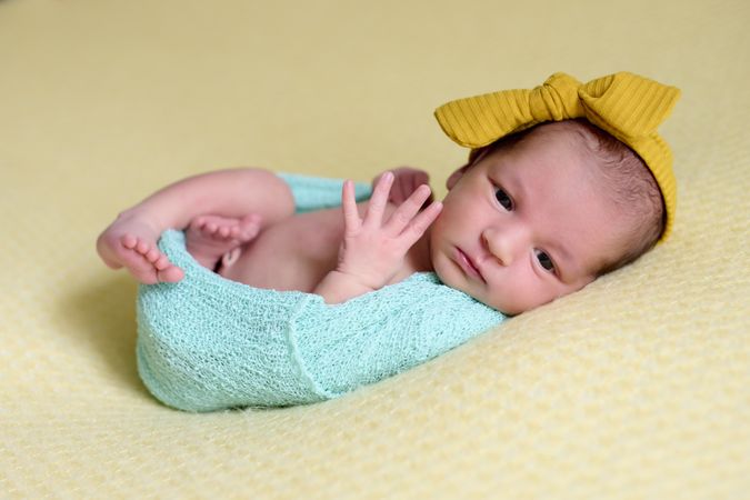 Close-up of a newborn baby girl, inside a ball of blue wool on a yellow blanket with a yellow bow on her head