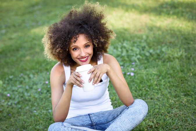 Black woman with afro hairstyle sitting cross legged in park with coffee