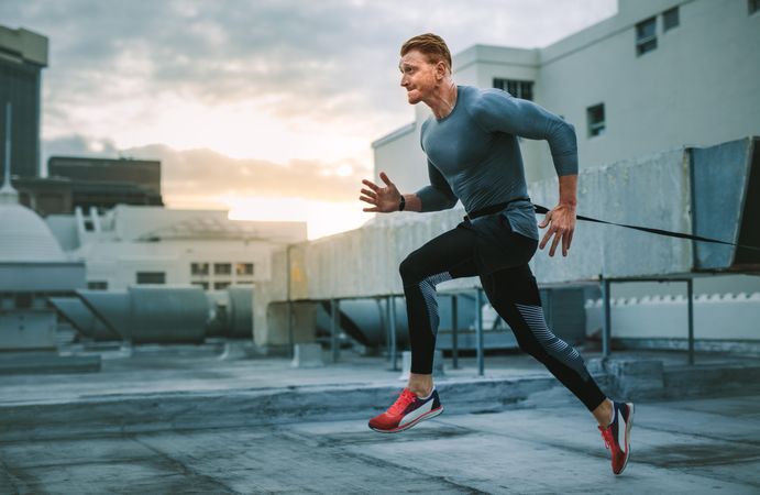 Fitness man running on terrace of a building with a resistance band to his waist