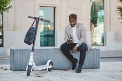 Black man sitting on bench in street with smartphone in shade o