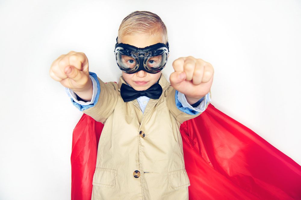 Serious blond boy wearing airplane goggles and cape with both fists out