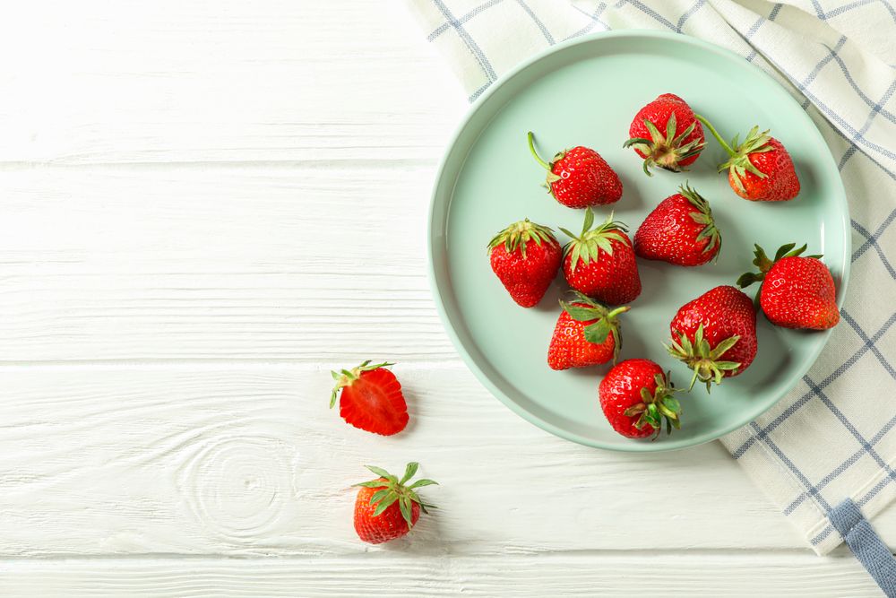 Plate with fresh strawberries and kitchen towel on bright wooden background, top view and space for text