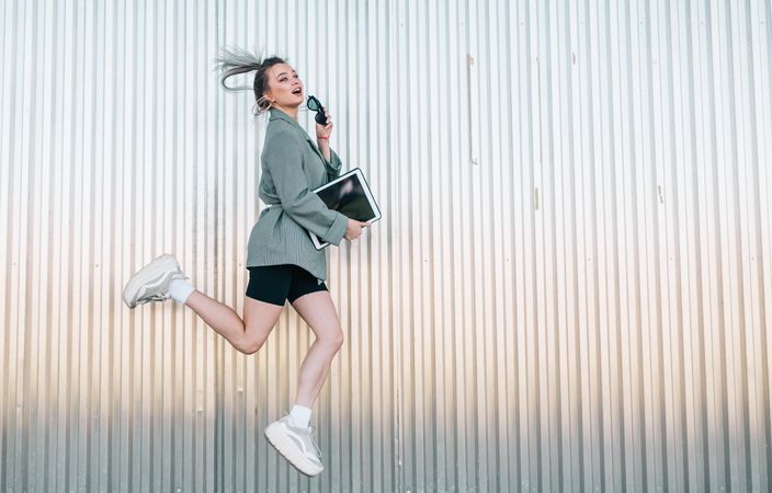 Woman with tablet in stylish suit jumping nearby wall holding sunglasses