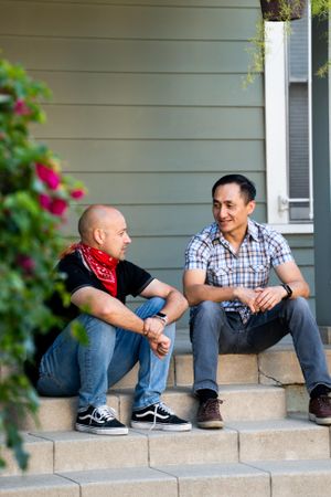Two men sitting on porch in front of their house talking and smiling