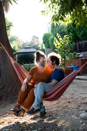 Young couple sitting in hammock with masks smiling at each other