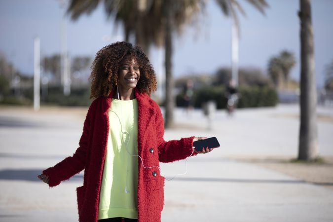 Smiling female in colorful clothes listening to music on phone standing on street with palm trees