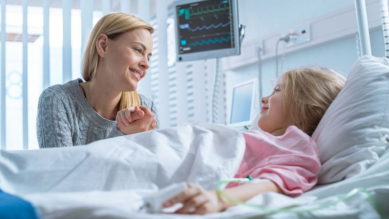Cute Little Girl Lies on a Bed in the Children's Hospital, Her Mother Sits Beside, They Talk