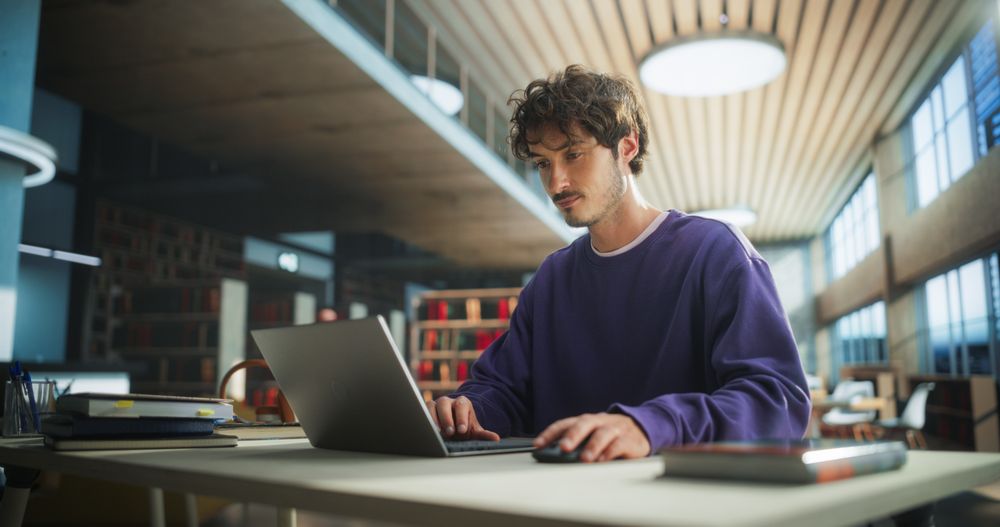 Student Studying in a Modern Library. Young Man Thinking and Problem Solving School Exercises. Male Using Laptop Computer to Work on a University Research Project Online