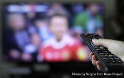Person holding remote control with people in the background on TV screen 4MvkGb