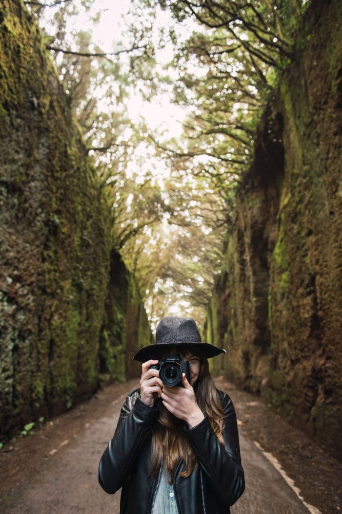 Young stylish woman taking photo on camera on way between high walls and trees