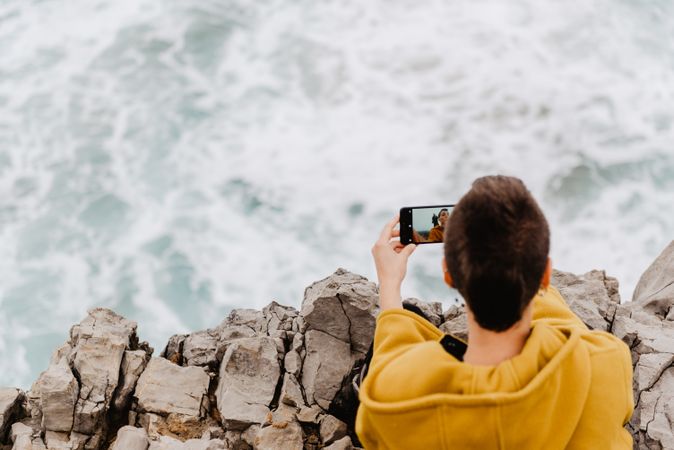 Woman in yellow hoodie taking selfie on smartphone on rocky shore