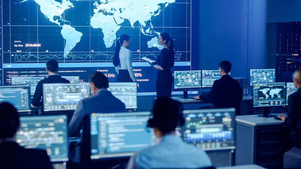 Diverse Team in a High-tech Control Room Analyzes Global Data on Multiple Screens, Ensuring Cybersecurity and Efficient Operations.