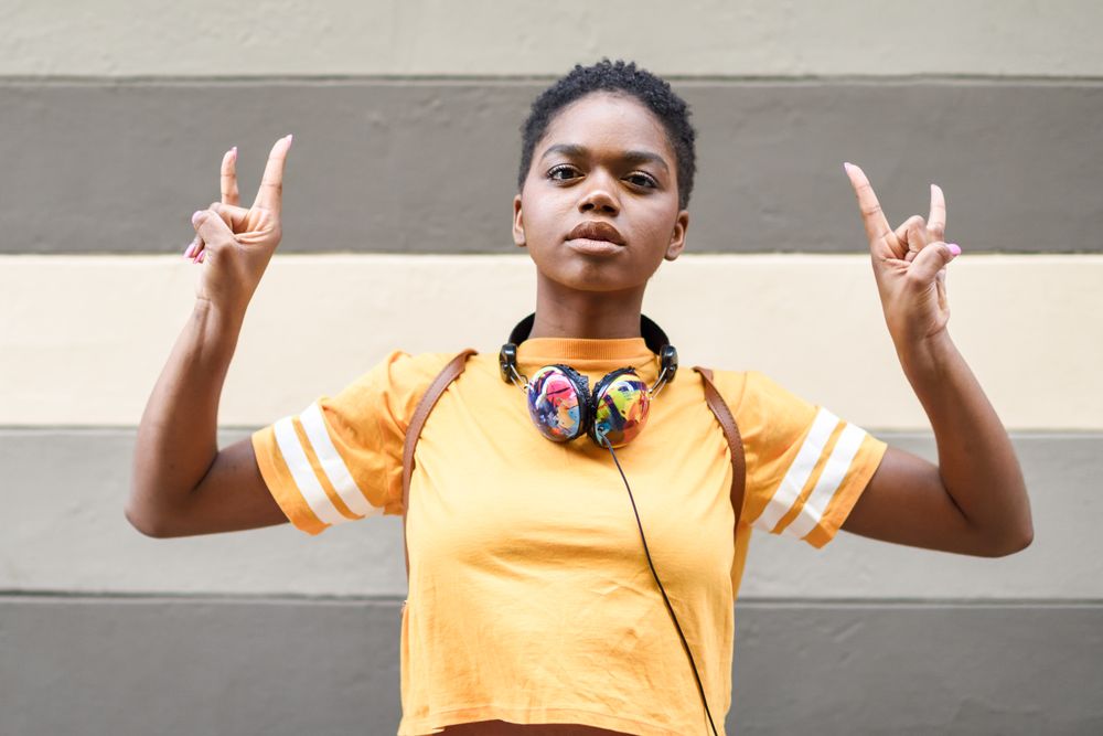 Black woman making metal hand sign with headphones in front of grey striped wall