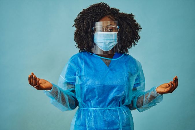 Serious Black female medical professional in surgical PPE, eye mask and shield, with arms open