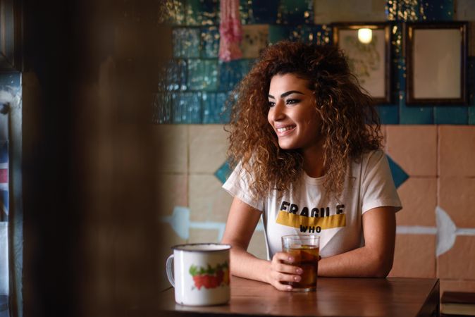 Arab woman in casual clothes smiling into the distance with soda at restaurant table