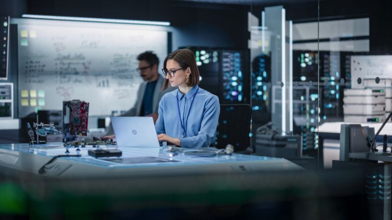 Portrait of Female Specialist Using Laptop Computer to Improve Electronic Circuit Board in Modern Office. Professional Engineer Thinking and Typing Ideas for Artificial Intelligence Project