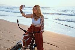 Cheerful woman taking selfie on smartphone while resting with bicycle at sunny shore