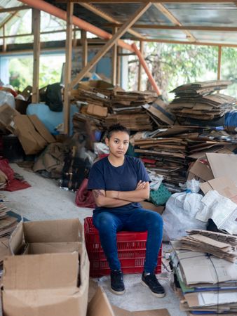 Young Hispanic Woman Determined Among Cardboard for Recycling in Warehouse. Serious Portrait