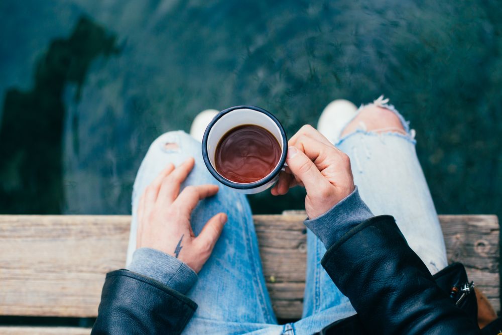 Man with tattoo on hand holding enamel coffee cup on pier