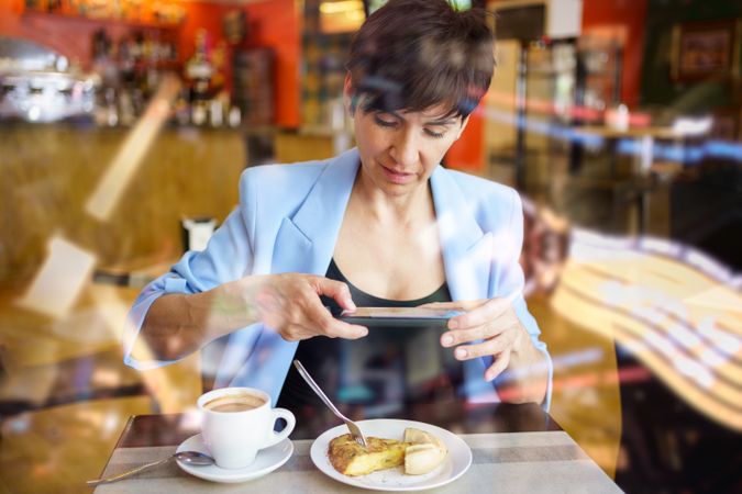 Female seen behind glass in trendy blue jacket sitting at table taking picture of slice of cake in cafe