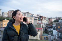 Joyful Asian female tourist in warm wear calling on smartphone at big old city
