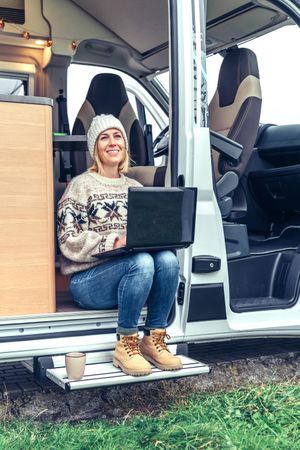 Female in sweater working remotely sitting on step of van while on a road trip, vertical
