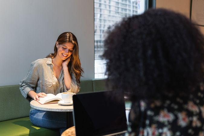 Woman reading book against crop ethnic girlfriend in cafe