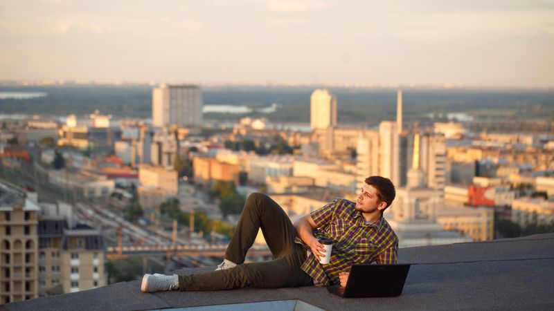 Man lying back on roof over looking city working on laptop with takeaway drink
