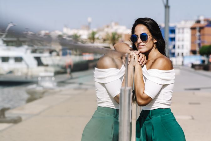 Side view of dreamy young woman with sunglasses leaning on fence and looking away on harbor dock
