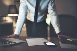 Man in shirt and tie leaning over desk with documents, cell phone and laptop
