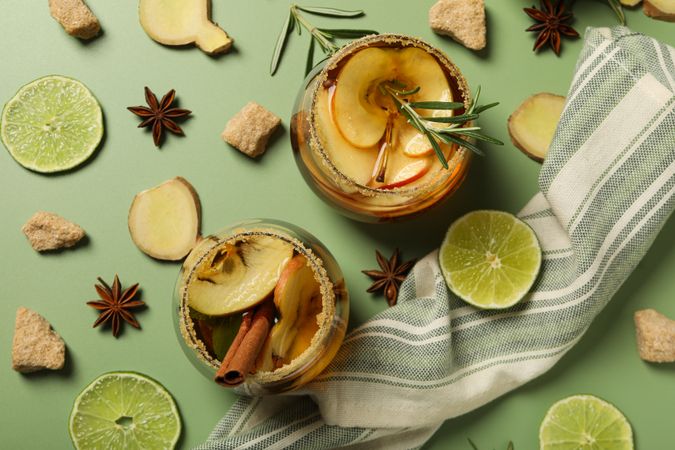 Glasses with cider, apple, lime and ginger pieces on green background, top view