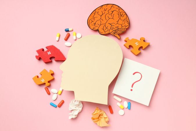 Paper cut out of side view of head with medications and puzzle pieces on pink background