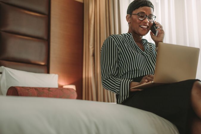 Smartly dressed woman talking on mobile phone while sitting in the hotel room using laptop