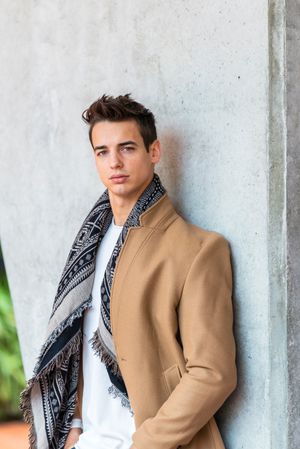 Serious man wearing camel coat and scarf looking at camera against cement wall