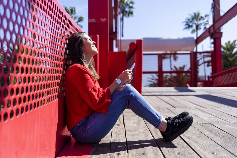 Side view of happy woman in red sitting talking on mobile phone outdoors on a bright day