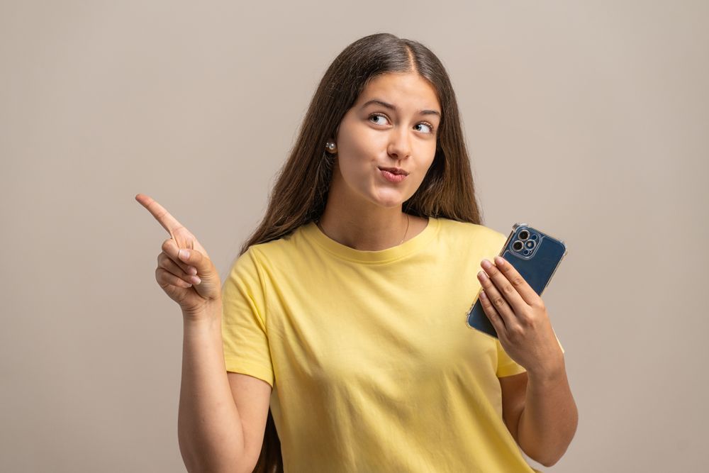 Thoughtful young woman holding a phone and pointing, considering an option.
